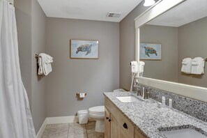 Un baño con ducha, inodoro, lavamanos y un espejo grande. Hay toallas colgadas en la pared y un cuadro con una tortuga.
