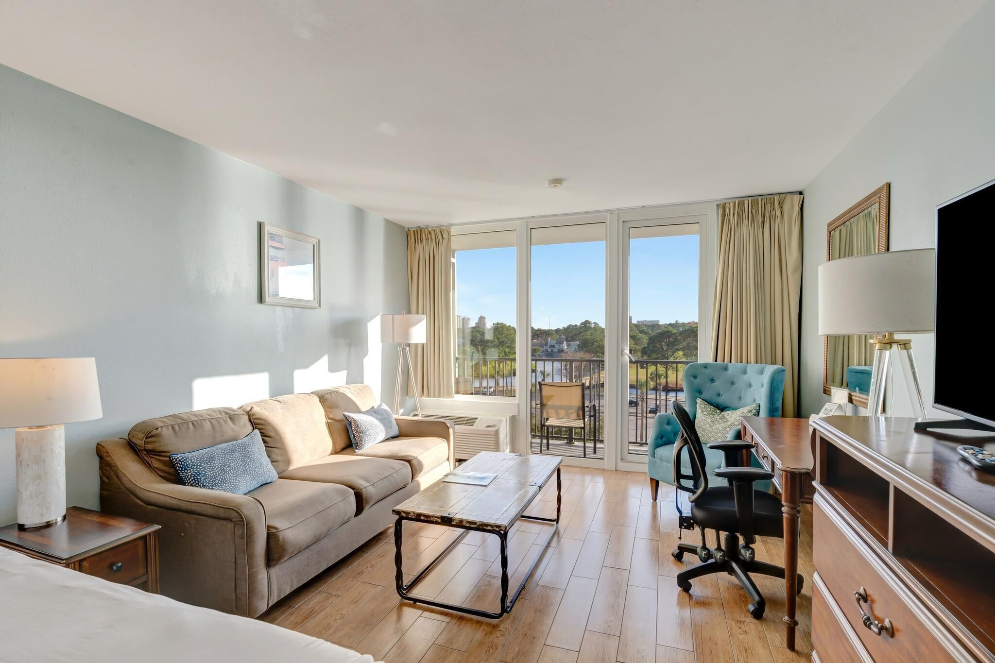 A hotel room with a sofa, a desk, a chair, a TV, and a balcony with a view.