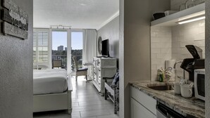 Habitación de hotel compacta con cama, mesa de comedor, sofá, televisor, kitchenette con lavabo y microondas, y balcón con vista a la ciudad.