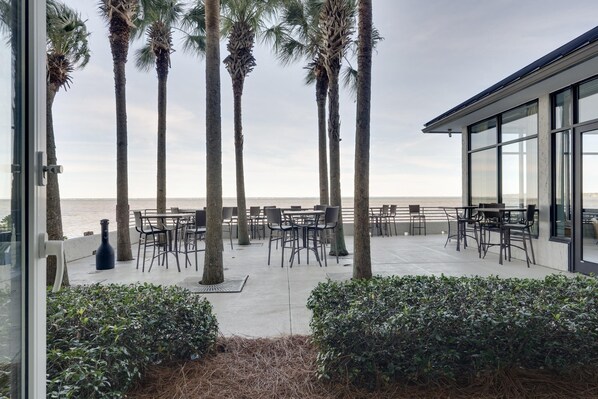 An outdoor patio with tables and chairs, palm trees, and a view of the sea. - Seaside Serenity - Coastal Studio Escape with King Bed for Two! at Sandestin Golf and Beach Resort! (Miramar Beach)