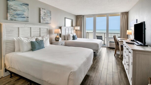 A hotel room with two beds, a desk, a TV, and a balcony with a view of the sea.