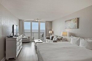 A hotel room with a large bed, a desk with a television, a chair, a table, and a balcony with a view.