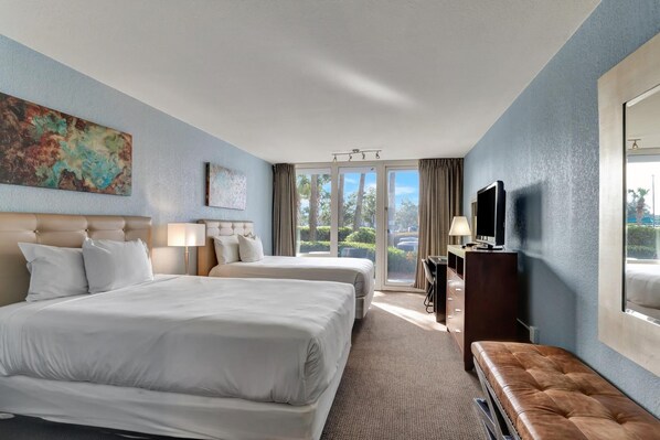 A hotel room with two beds, a television, a desk, and a large window offering a view of trees and a parking area.