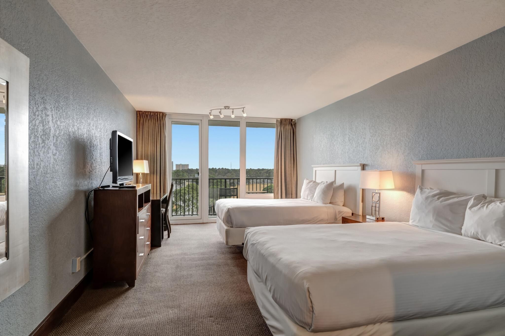 A hotel room with two beds, a TV, a desk, and a balcony with a view.