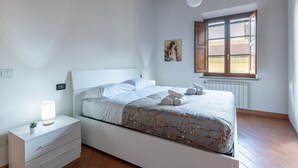 Un dormitorio con cama, mesita de noche, lámpara, ventana y una obra de arte.