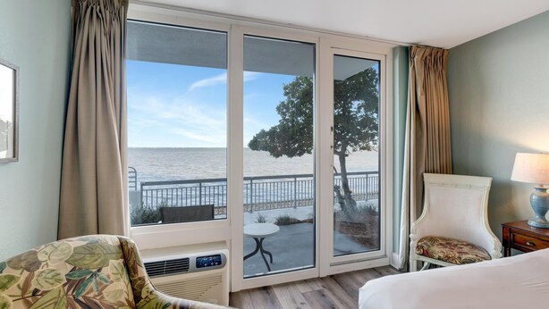 Een hotelkamer met een grote schuifglazen deur naar de zee, een bed, een stoel en een klein tafeltje.