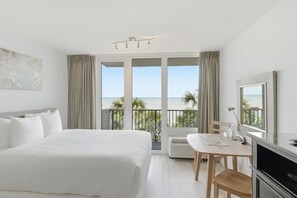 A hotel room with a bed, a desk, a chair, a TV, and a balcony with a view of the sea.