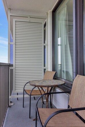 Een balkon met een tafel en twee stoelen, een raam met zonwering en een metalen hekwerk.