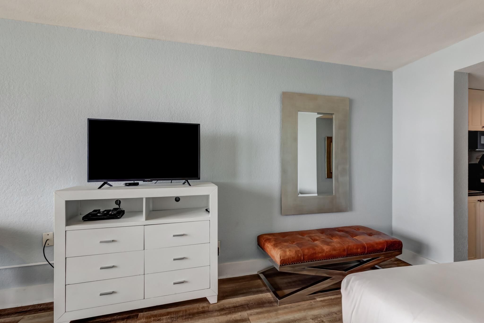 Ein Hotelzimmer mit einem weißen Schminktisch, einem Fernseher darauf, einem braunen Ledersitzhocker und einem großen Spiegel an der Wand.