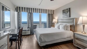 Une chambre d’hôtel avec un grand lit, un bureau et une chaise. Les fenêtres sont dotées de rideaux, et on peut admirer la vue sur la mer.