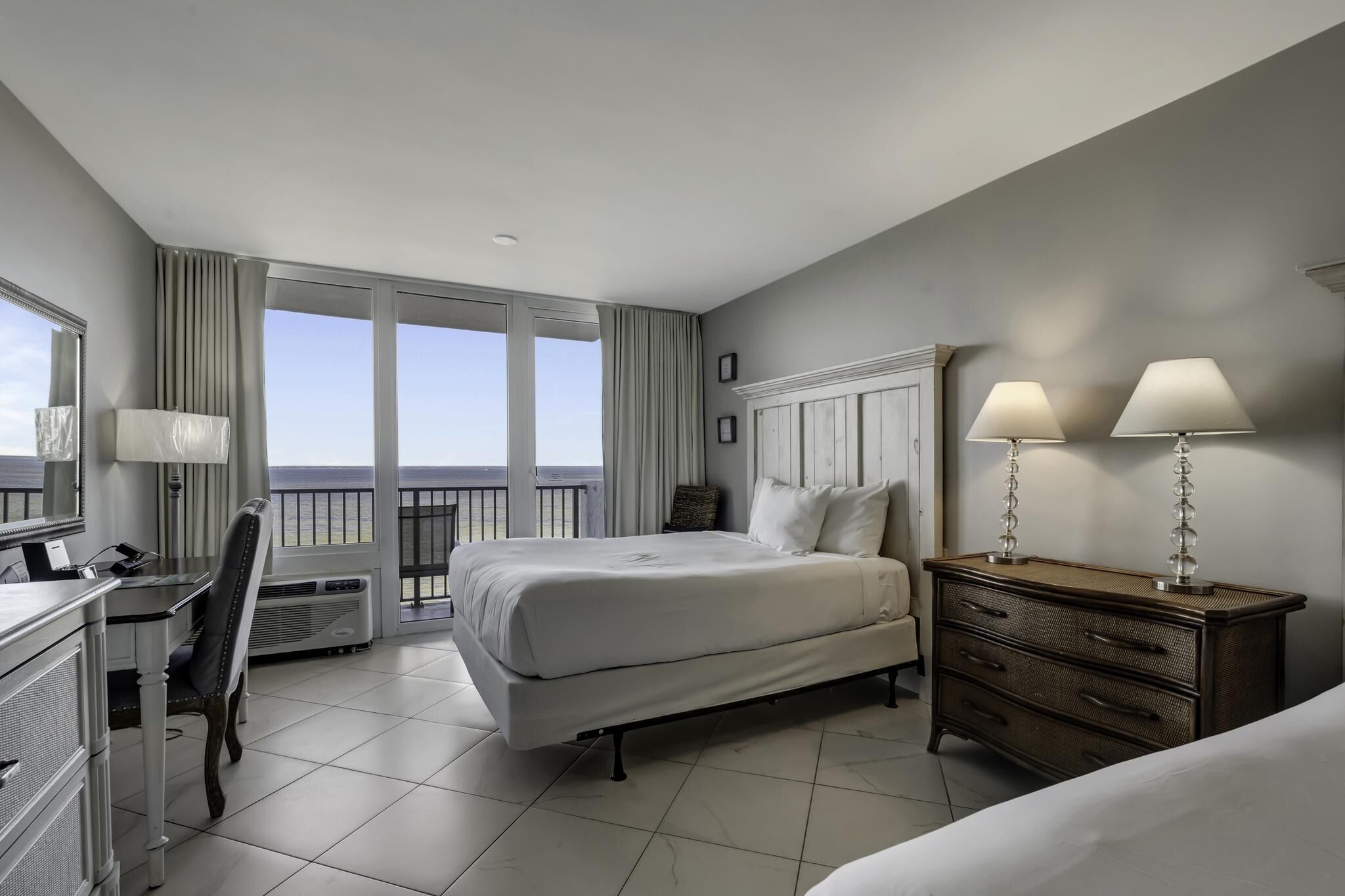 A hotel room with a bed, a desk, a chair, a dresser, and a balcony with a view.