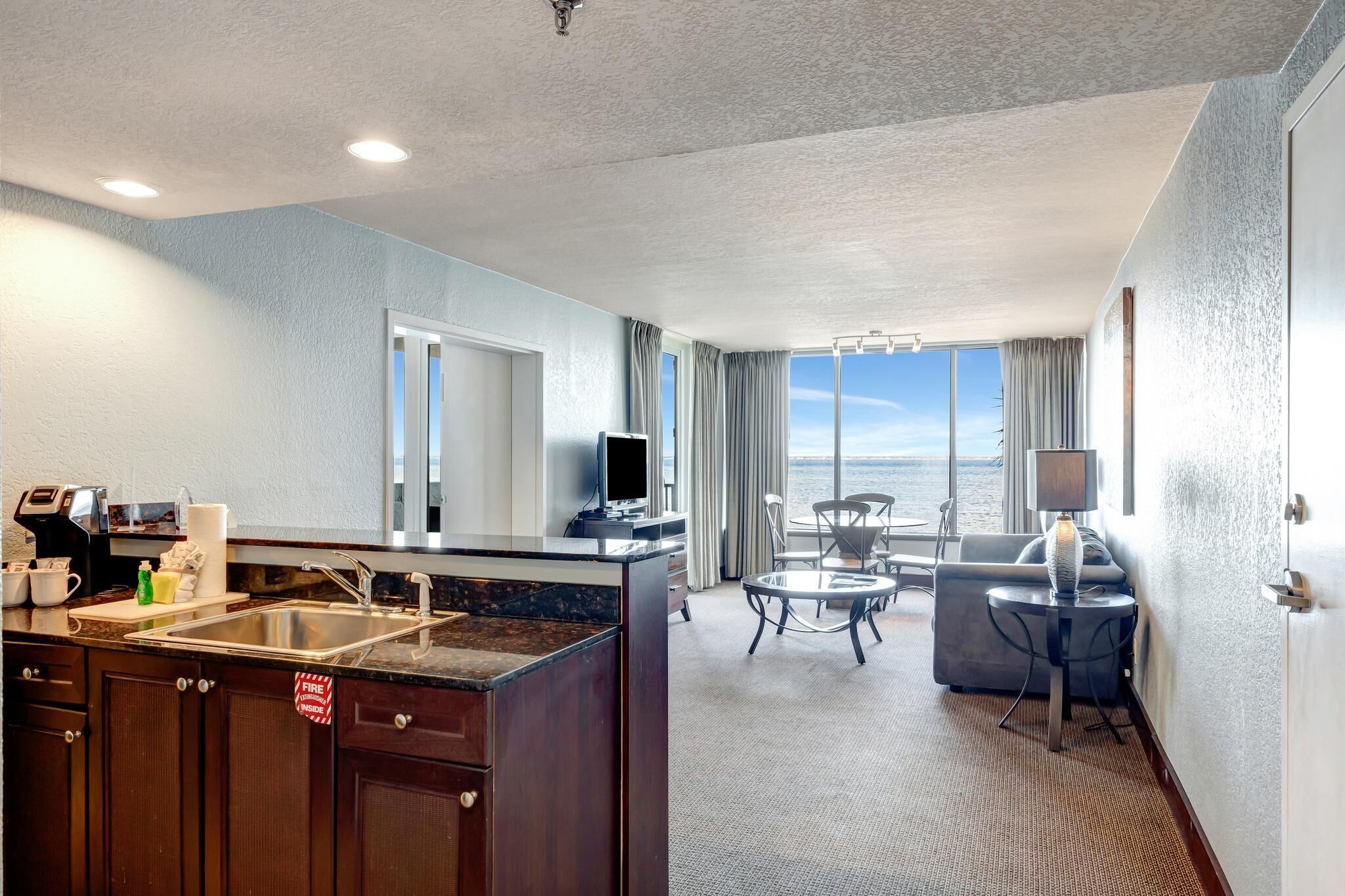 A hotel room with a kitchen area, a dining table with chairs, a sofa, and a large window offering a view of the ocean.