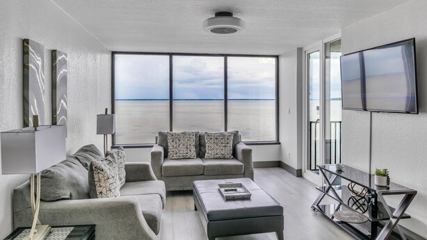 Een moderne woonkamer met een groot raam met uitzicht op zee, een bank, een salontafel en een aan de muur bevestigde televisie.