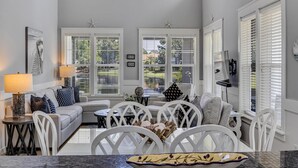 Un soggiorno luminoso e ben illuminato con un divano bianco, poltrone abbinate e un tavolo da pranzo. Sono presenti ampie finestre con persiane, decorazioni a stella marina e vista sul verde all'esterno.