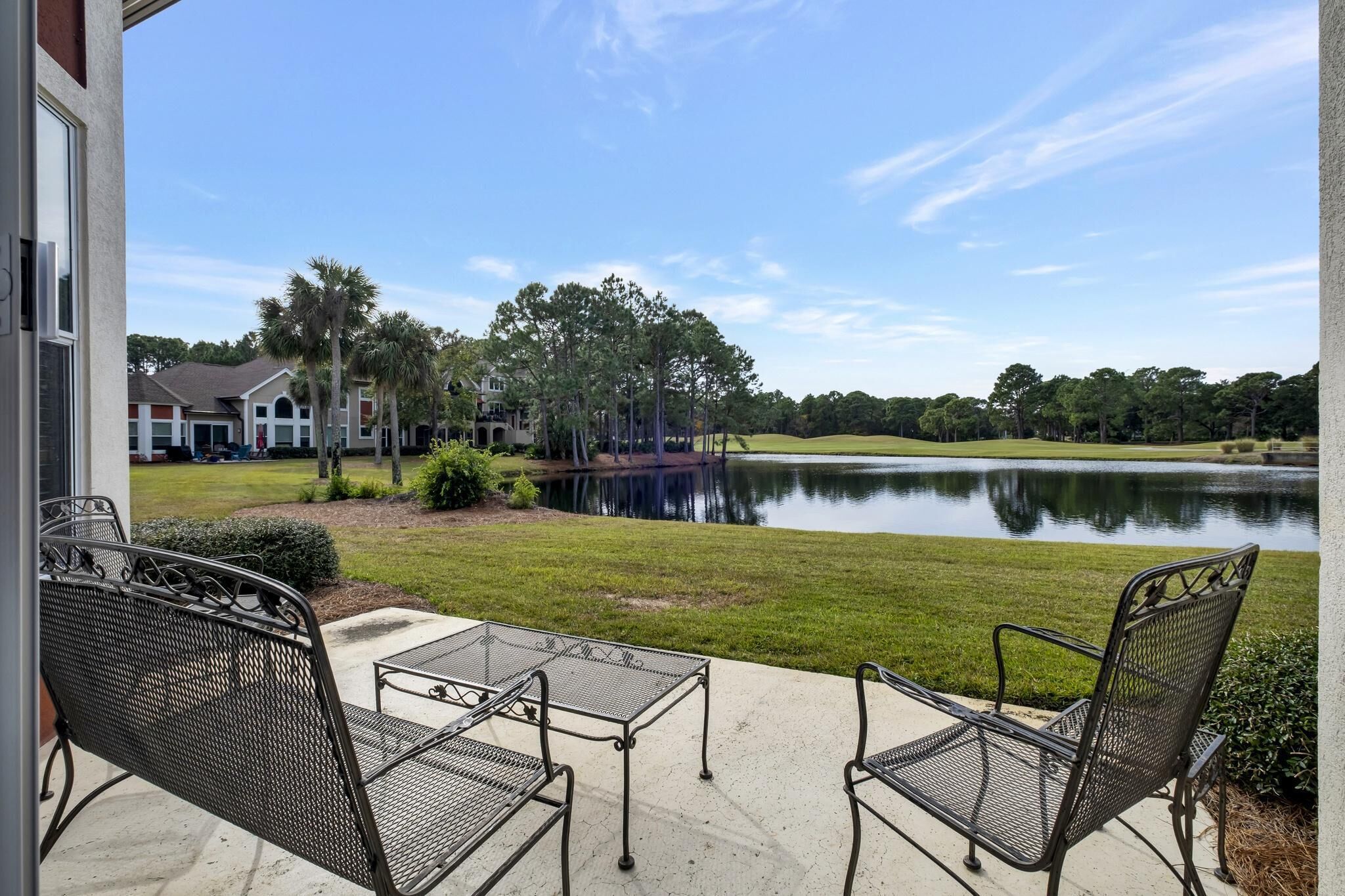 A patio with metal chairs and a table overlooking a lake, surrounded by houses and palm trees.
