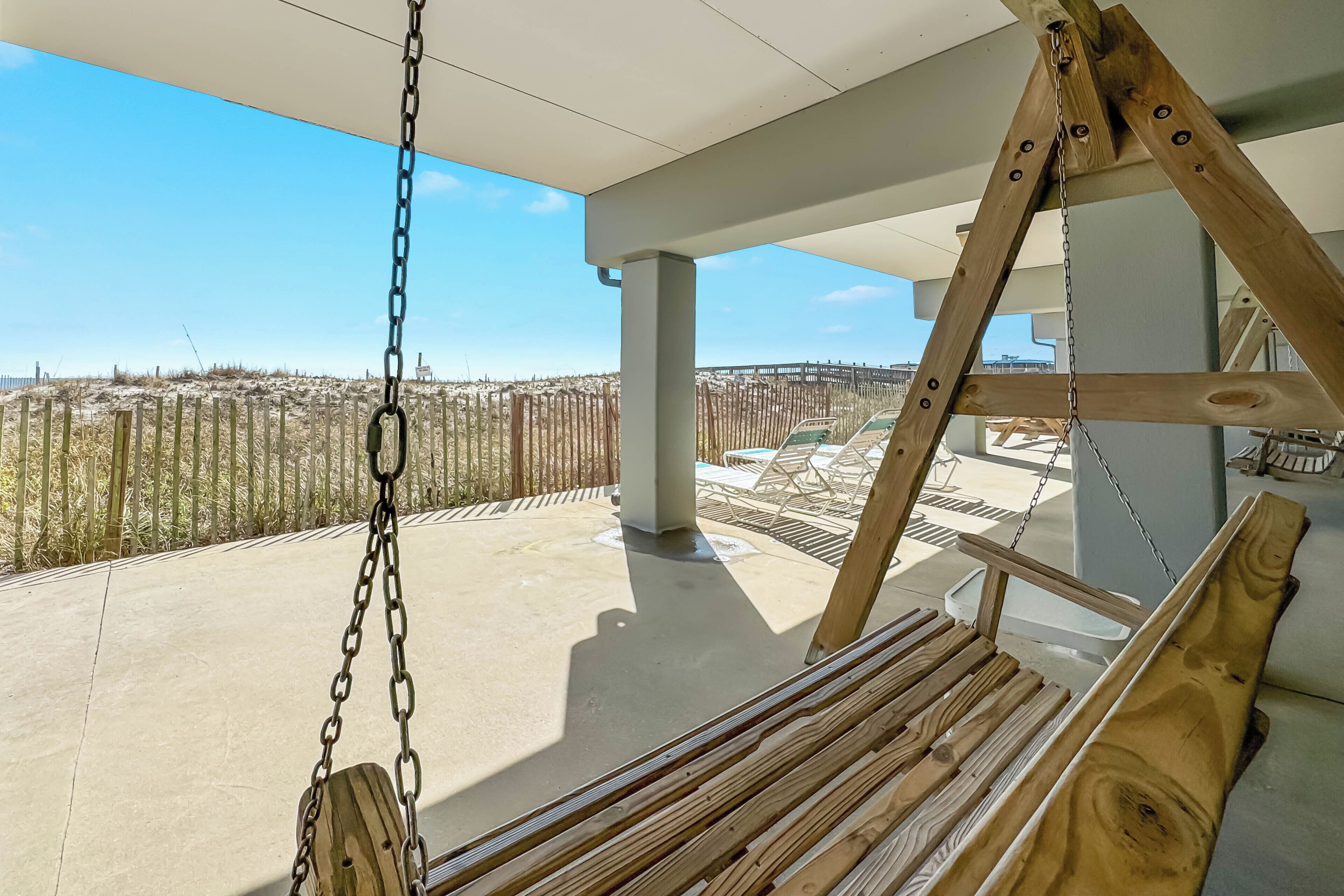 A wooden swing suspended from a ceiling, overlooking a beach with a wooden fence and lounge chairs.