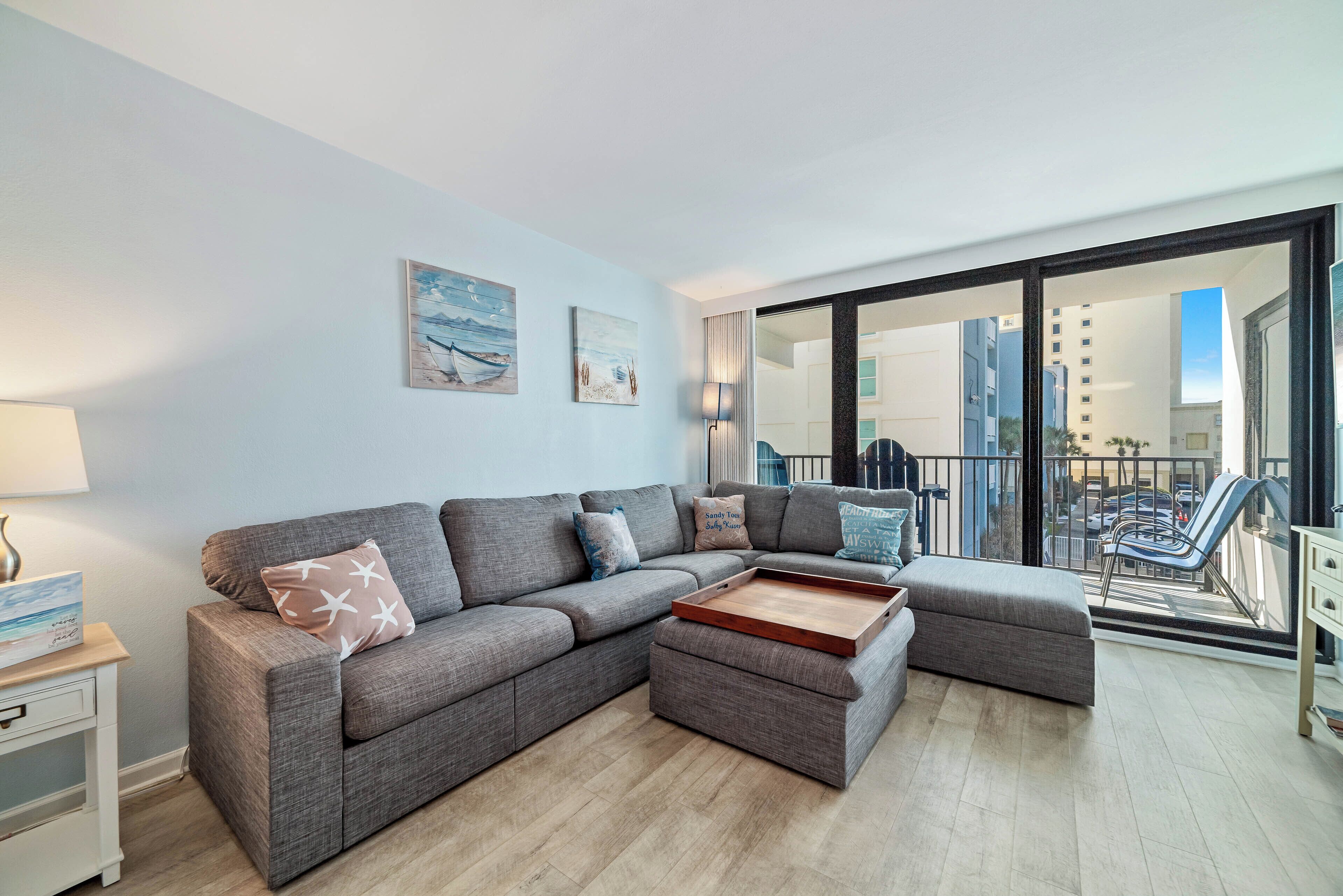 Ein modernes Wohnzimmer mit einem grauen Ecksofa, einem Holztisch und einem Balkon mit Blick auf die Stadt.