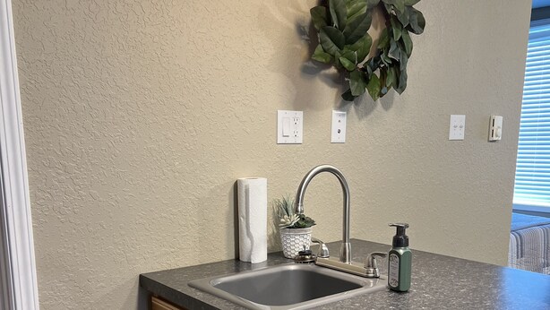 A kitchen with a granite countertop, wooden cabinets, a stainless steel sink, and a wall-mounted light fixture. - The Petite Perch (Alden)