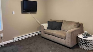 A room with a beige wall, a ceiling fan, a flat-screen TV mounted on the wall, a beige sofa with cushions, a small round side table, and a white radiator. - The Petite Perch (Alden)
