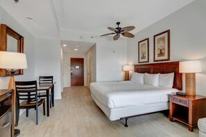 A hotel room with a bed, two bedside tables with lamps, a ceiling fan, a desk with a chair, and a door leading to another room.