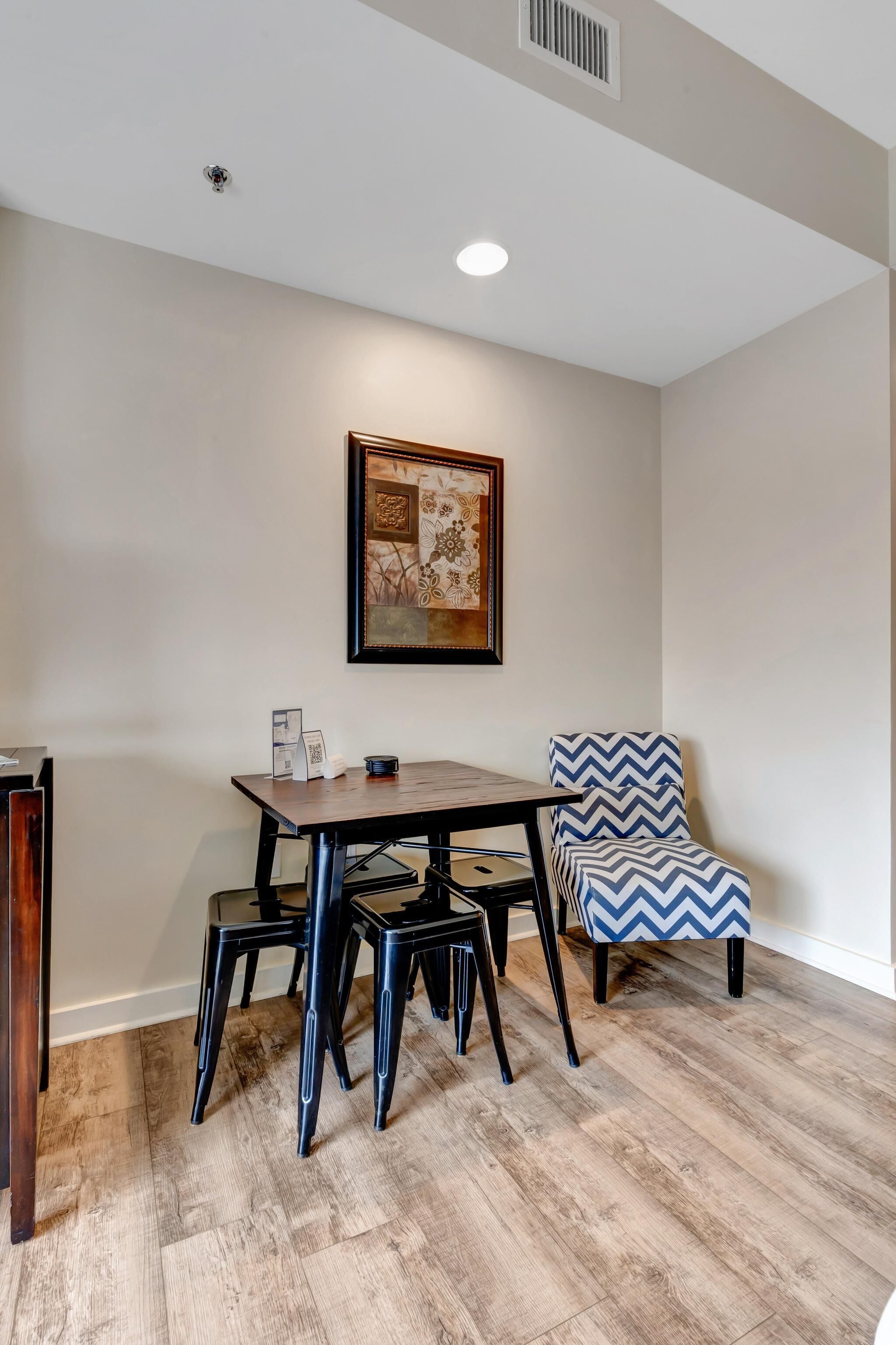 A dining area with a table and chairs, a framed artwork on the wall, and a vent on the ceiling.