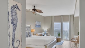 Ein Hotelzimmer mit Bett, Deckenventilator, Fenster mit Vorhängen, einem Balkon und einem Kunstwerk mit Seepferdchen an der Wand.