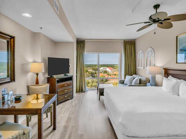 A hotel room with a bed, a TV, a desk, a chair, a ceiling fan, and a large window with a view.