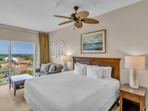 A hotel room with a large bed, a ceiling fan, a view of houses, and a painting on the wall.