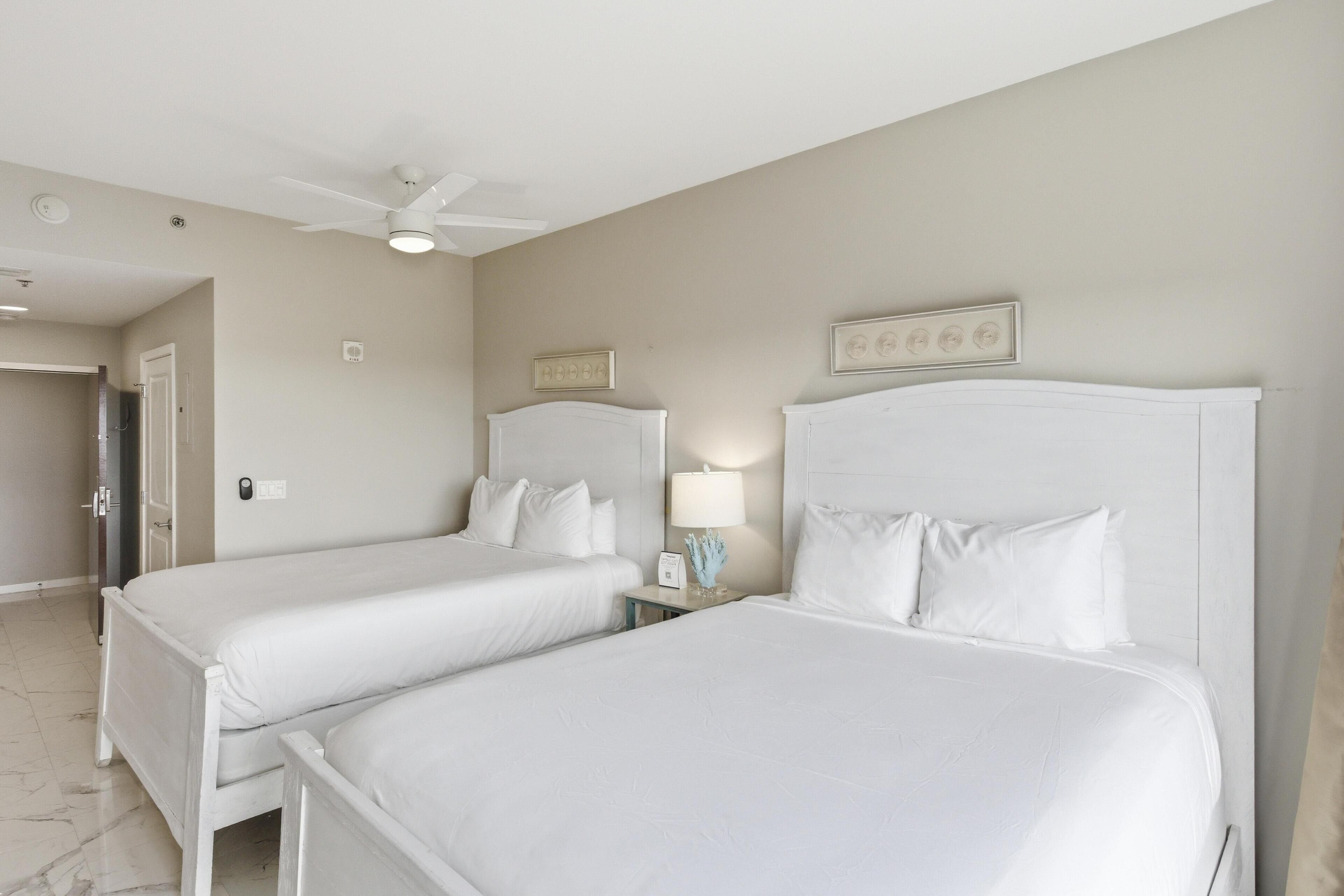 Une chambre d’hôtel avec deux lits, un ventilateur de plafond, une table de chevet avec une lampe et une porte donnant sur une autre pièce.