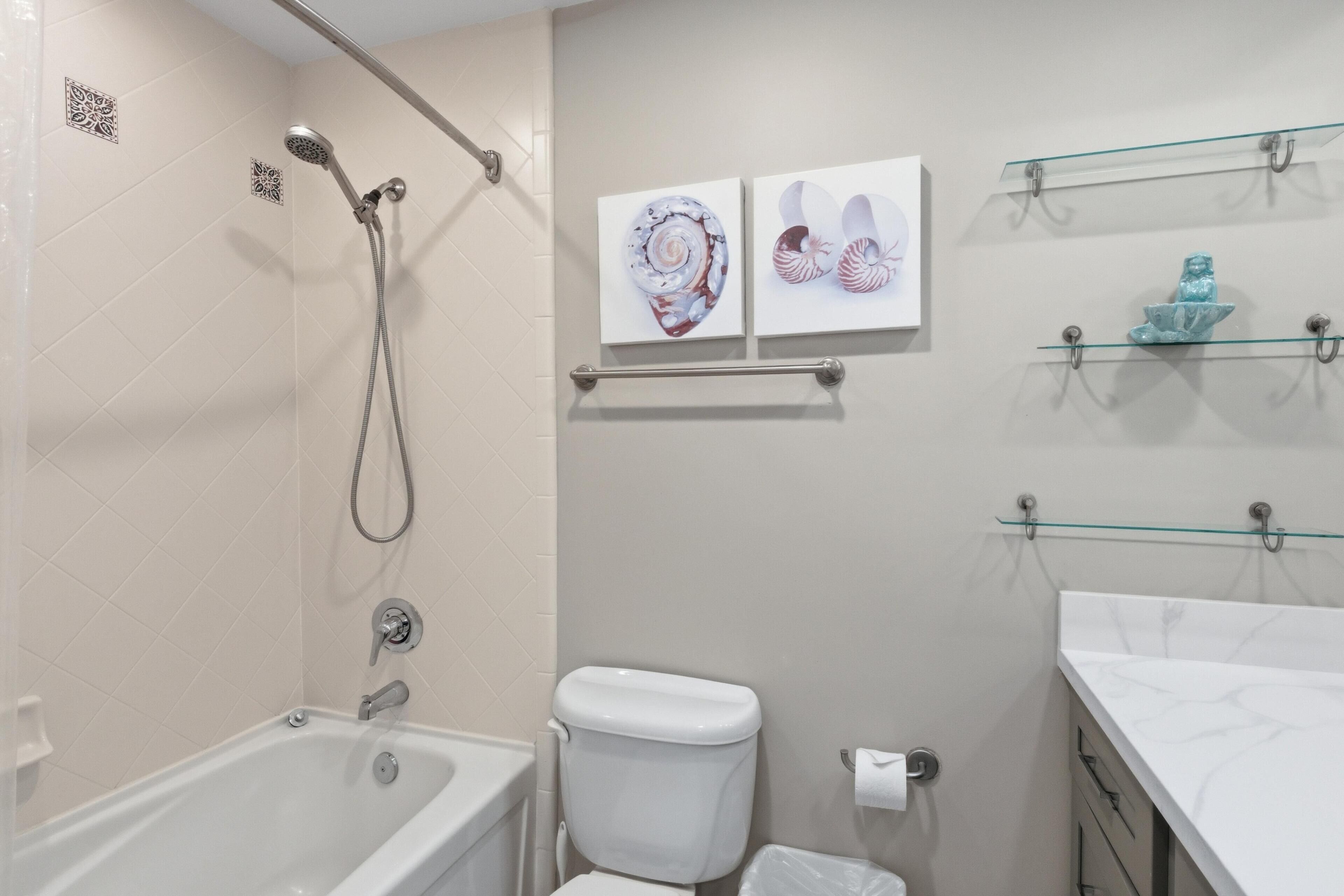 Een badkamer met een bad, toilet en douche. Twee ingelijste kunstwerken hangen aan de muur en er is een glazen plankje met een decoratief object.