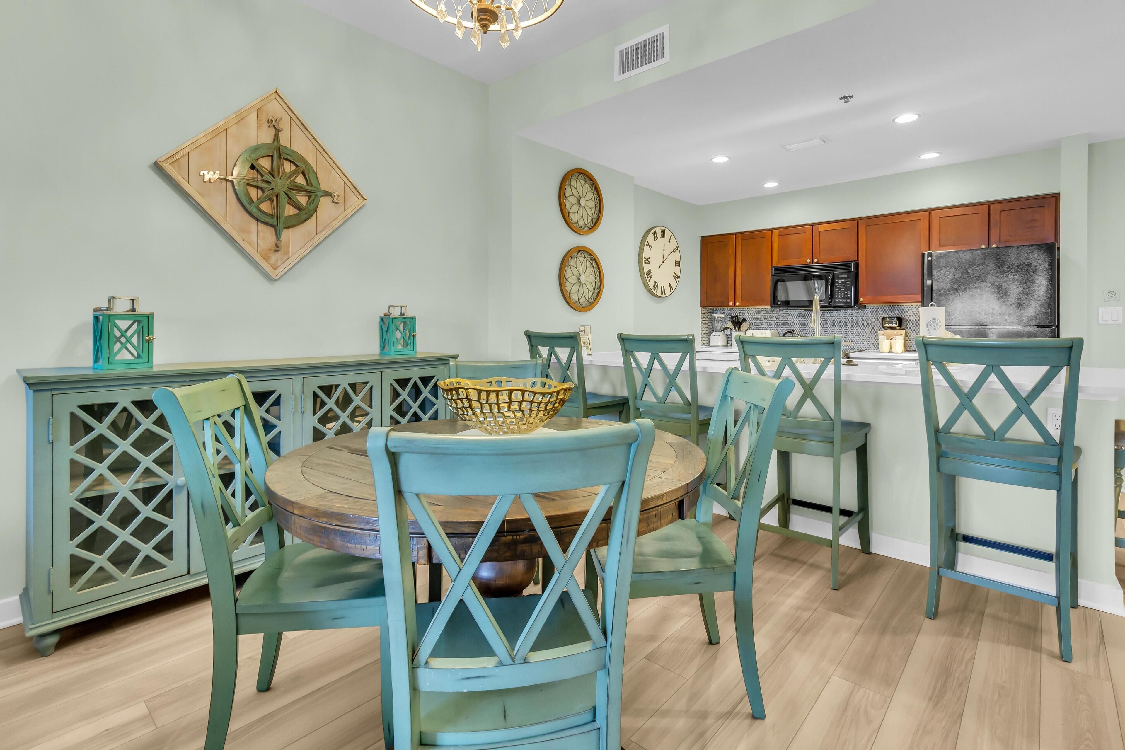Un comedor con sillas color turquesa y una mesa de madera, una cocina con gabinetes y electrodomésticos de madera, y relojes decorativos en la pared.