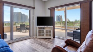 Un'ampia zona giorno con televisione a schermo piatto su un mobile bianco, grandi porte scorrevoli in vetro che si aprono su un balcone e vista su edifici moderni.