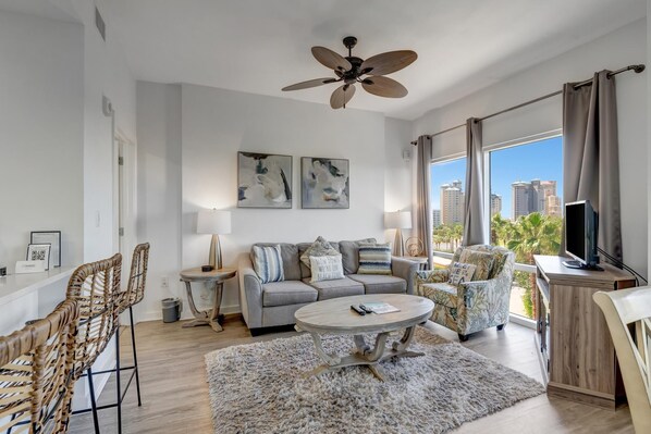 Ein modernes Wohnzimmer mit einem grauen Sofa, einem runden Couchtisch, Korbstühlen und einem Deckenventilator, sowie einem großen Fenster mit Blick auf die Stadt.