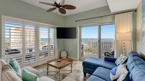 Ein Wohnzimmer mit einem blauen Sofa, einem Holztisch und einem Deckenventilator. Ein Fernseher ist an der Wand montiert und große Fenster bieten Blick auf Gebäude und den Himmel.
