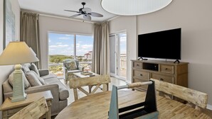 Una sala de estar moderna con un sofá, una mesa de comedor de madera, un televisor y un balcón con vistas a edificios.