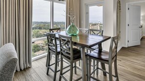 A dining area with a wooden table, chairs, and a vase. Large windows offer a view of the city. - Paradise Found: Luau Condo with Lagoon Pool & More (Miramar Beach)