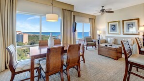 A hotel room with a dining area, a sofa set, a ceiling fan, and large windows offering a city view.