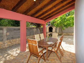 Une terrasse extérieure avec un sol en pierre, une table et des chaises en bois, un mur en pierre et un four en briques en arrière-plan.