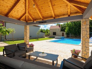 Une terrasse extérieure avec un sol en pierre, une piscine et un espace salon couvert comprenant des canapés et une table.