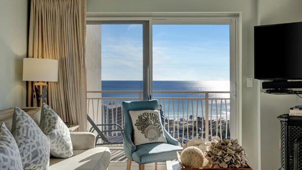 A room with a view of the sea, a sofa with patterned cushions, a blue armchair with a decorative pillow, a lamp, and a television. - Sandestin Escape: Spacious 2BR with Ocean Views, Poolside Bar, Resort Perks & More (Miramar Beach)