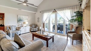 A spacious living room with a ceiling fan, a large window with curtains, a dining area with a table and chairs, and a view of the outdoors. - 3BR/3BA Golf Course View Retreat, Sleeps 6 (Miramar Beach)