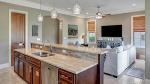 Una cocina moderna con una isla grande, electrodomésticos integrados, iluminación colgante y un comedor con sofá y televisor.