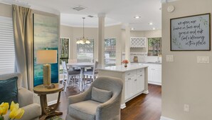 A modern living room with a grey armchair, a dining area with a table and chairs, and a kitchen with white cabinets. - Charming 3BR/3BA Sandestin Home: Spacious Comfort, Golf Cart, Sleeps 6 (Miramar Beach)