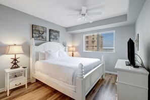 Ein Schlafzimmer mit einem weißen Bett, zwei Nachttischlampen, einer Lampenform von einem Seestern, einem Deckenventilator, einem Fenster mit Jalousien und einem Schminktisch mit Fernseher.