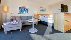 Quarto de hotel com sofá, cama, mesa pequena, cômoda e duas luminárias fixadas na parede.