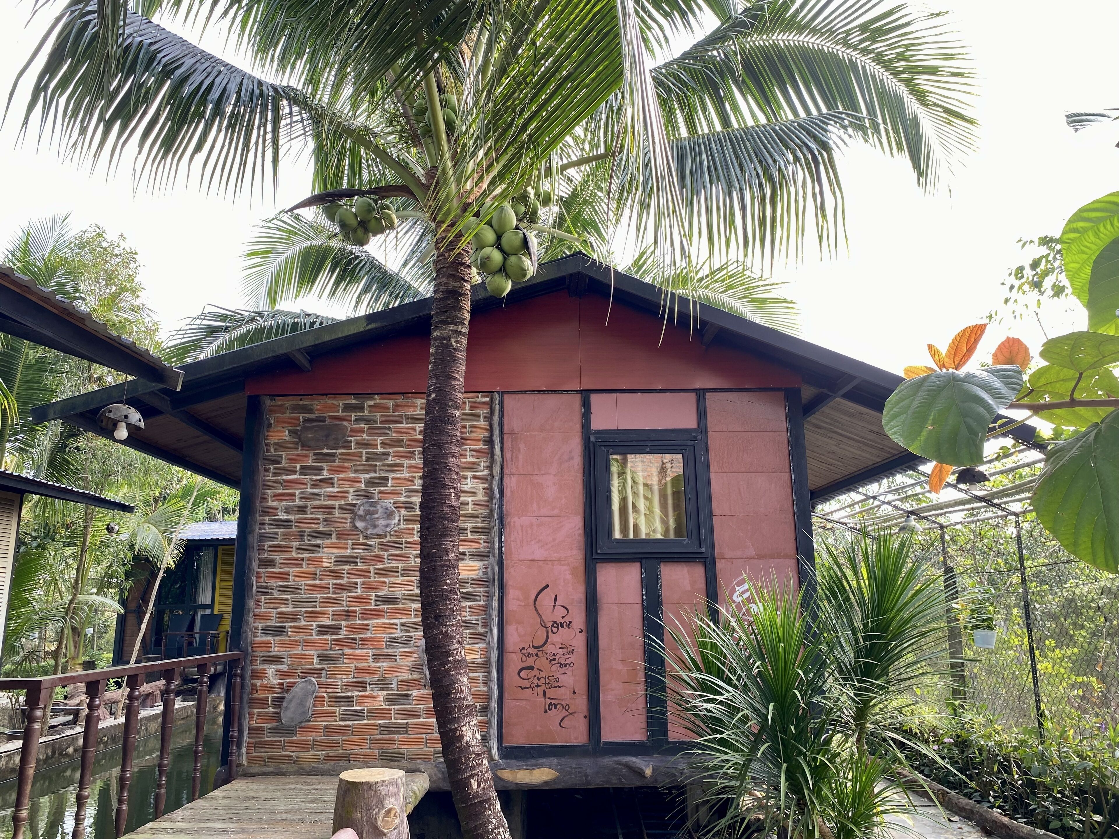 A small, rustic building with a brick wall and a wooden roof, surrounded by lush greenery and a palm tree.