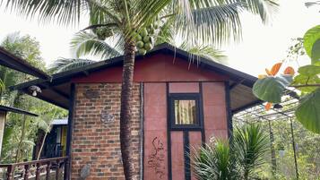 A small, rustic building with a brick wall and a wooden roof, surrounded by lush greenery and a palm tree.