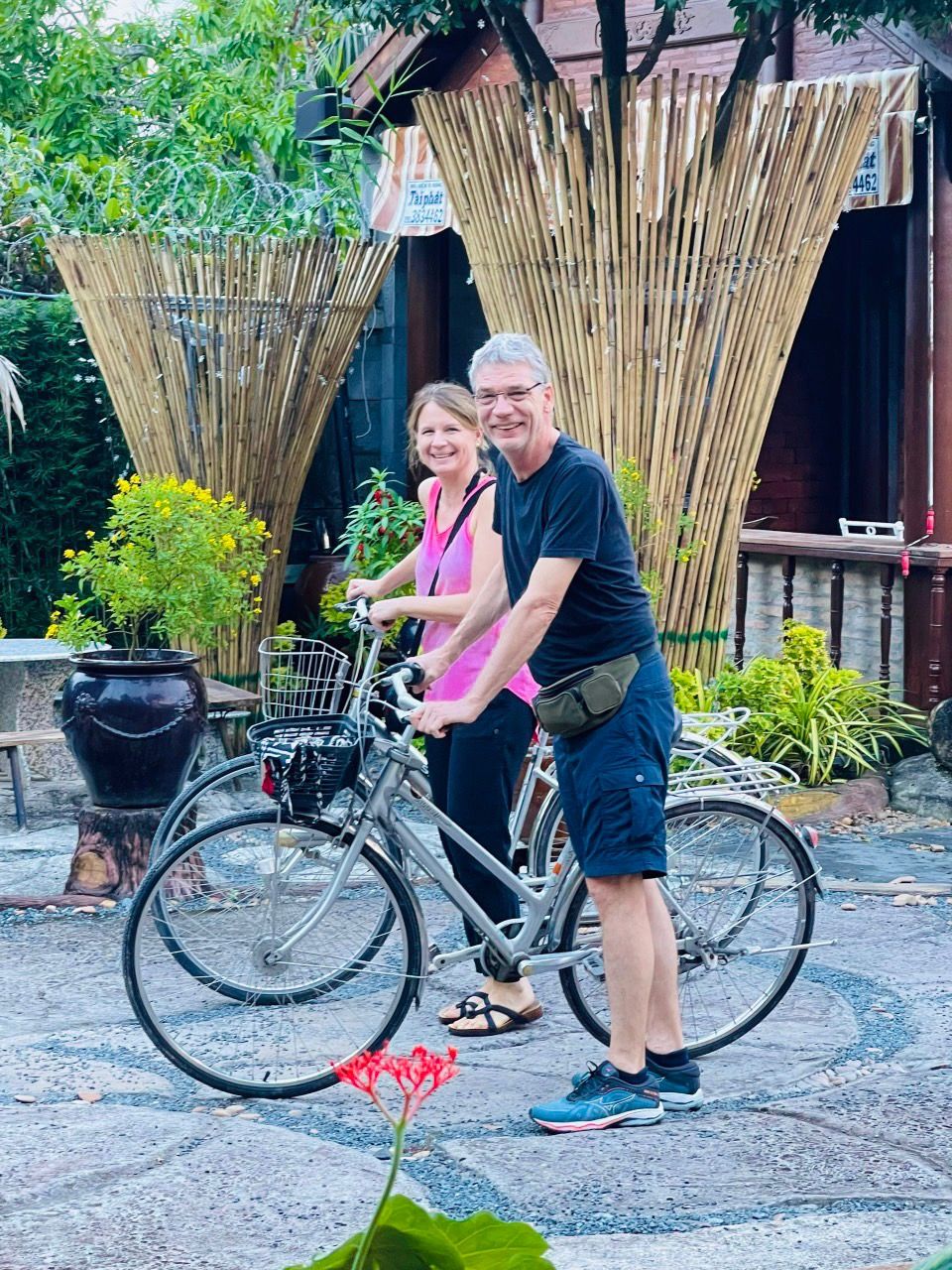 Una pareja de pie junto a una bicicleta con cesta, frente a una valla de bambú y un edificio con un letrero.