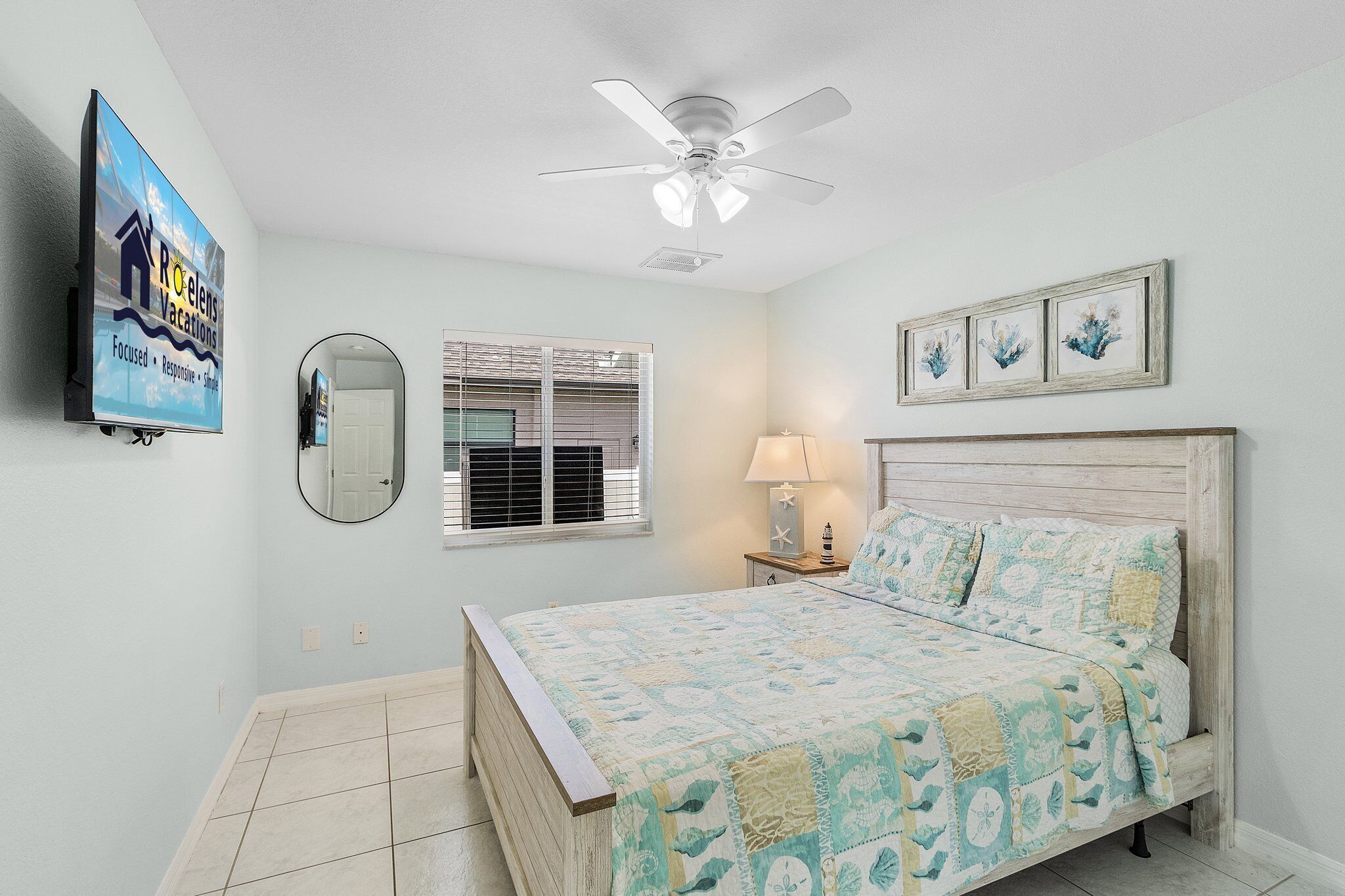 A bedroom with a bed, a ceiling fan, a TV, a window with blinds, a lamp, and framed artwork on the wall.
