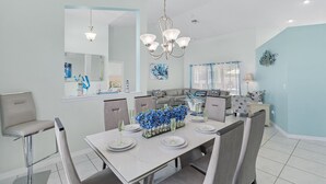 Un comedor moderno con una mesa blanca dispuesta para cuatro personas, rodeada de sillas grises. Un candelabro cuelga encima, y un centro de mesa floral azul decora la mesa.